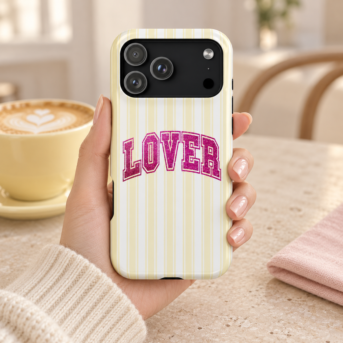 Lover Varsity MagSafe case, pink varsity LOVER lettering on pale yellow stripes, lifestyle photo in hand with coffee