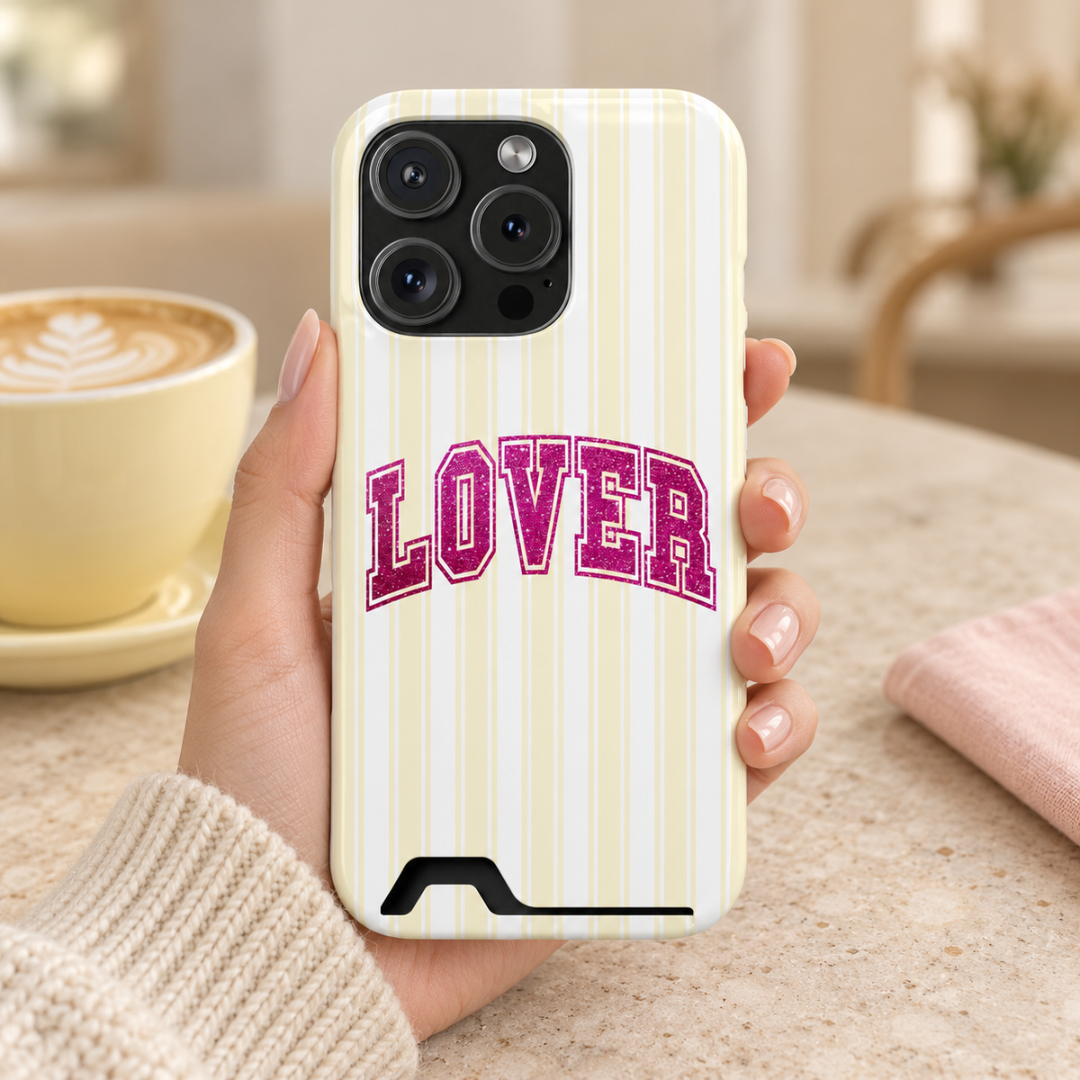 Lover Varsity phone case with card holder, pink varsity LOVER lettering on pale yellow stripes, lifestyle photo in hand with coffee